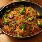 Gebratene Reisnudeln mit Sojasauce, serviert mit Broccoli, Karotten und Pilzen in einer Pfanne.