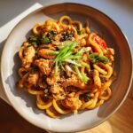 Gebratene Udon Nudeln mit Huhn, Gemüse und Sesam in einer Schale, garniert mit Frühlingszwiebeln.