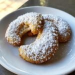 Two Vanillekipferl cookies on a white plate, generously dusted with powdered sugar.