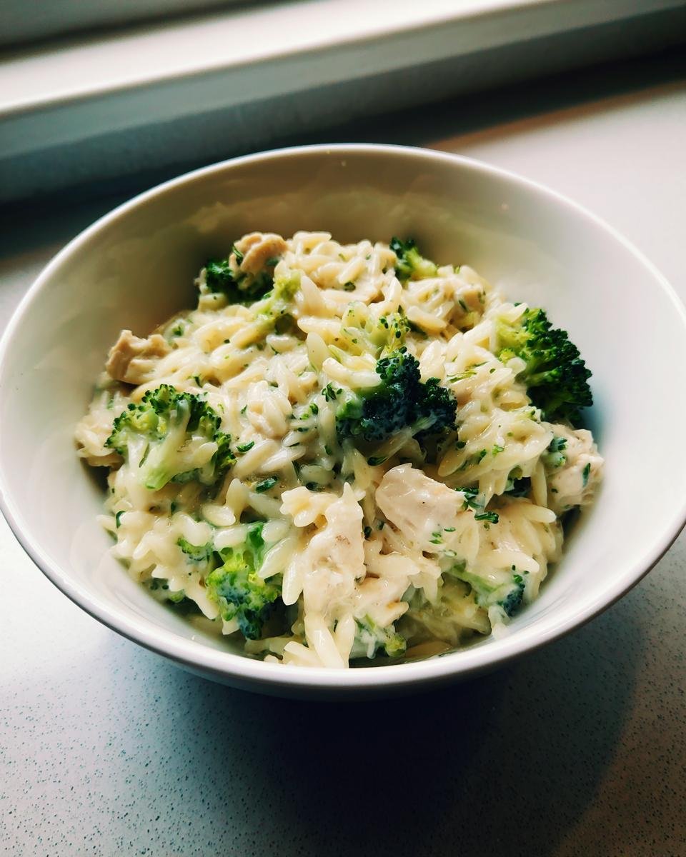 Eine weiße Schüssel gefüllt mit Cremige Hähnchen-Brokkoli-Orzo, bestehend aus cremiger Soße, Orzo-Nudeln, Hähnchenstücken und Brokkoli.