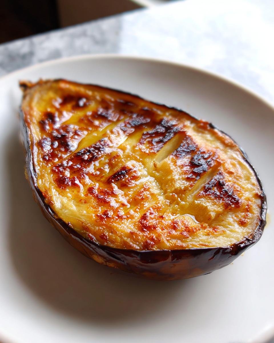 Nahaufnahme einer halbierten Goldbraune Ofen-Aubergine mit goldbraun überbackener Oberfläche auf einem weißen Teller.